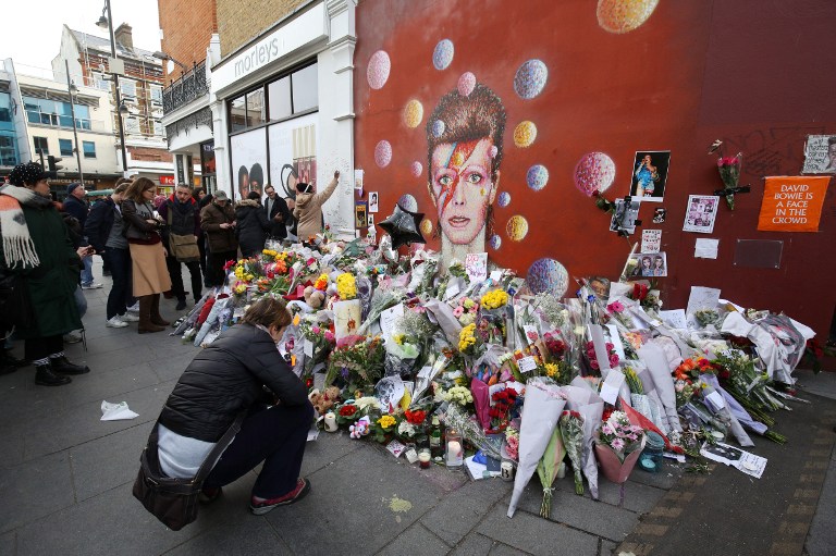 People gether around floral tributes in front of a mural of British singer David Bowie, painted by Australian street artist James Cochran, aka Jimmy C, in Brixton, south London, on January 13, 2016 two days after the announcement of Bowie's death. Music legend David Bowie was famously private during his lifetime -- and in death, as a string of questions about the circumstances of his passing remained unanswered. His official social media accounts had announced the shock news of his death at 69 on January 11, 2016: "David Bowie died peacefully today surrounded by his family after a courageous 18-month battle with cancer," adding a request for privacy for the grieving family. AFP PHOTO / JUSTIN TALLIS -- RESTRICTED TO EDITORIAL USE, MANDATORY MENTION OF THE ARTIST UPON PUBLICATION, TO ILLUSTRATE THE EVENT AS SPECIFIED IN THE CAPTION / AFP / JUSTIN TALLIS
