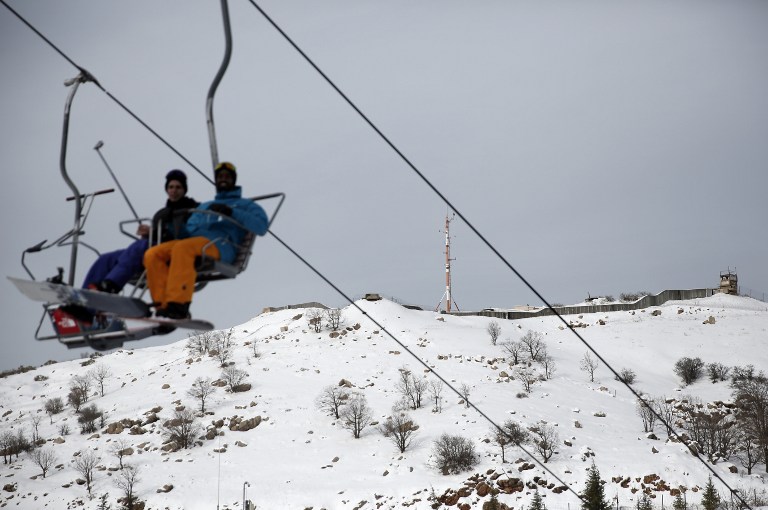 Skiers sit on a chairlift as an Israeli army base is seen in the background at the Israeli Mount Hermon ski resort, in the Israeli-occupied Golan Heights, on January 21, 2016. For Israelis, the Hermon range, straddling Lebanon and the Syrian and Israeli-held sectors of the Golan, is a highly strategic area under close surveillance. / AFP / THOMAS COEX / TO GO WITH AFP STORY BY DAPHNE ROUSSEAU