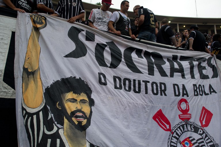Supporters of Corinthians stand next to a huge banner in homage of the late footballer Socrates, during the Brazilian Championship final date match against Palmeiras, at the Pacaembu stadium on December 04, 2011 in Sao Paulo, Brazil. Socrates -former captain of the national team and player of Corinthians- died earlier today at the age of 57 from an intestinal infection. AFP PHOTO / Yasuyoshi CHIBA / AFP PHOTO / YASUYOSHI CHIBA