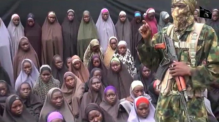 This video grab image created on August 14, 2016 taken from a video released on youtube purportedly by Islamist group Boko Haram showing what is claimed to be one of the groups fighters at an undisclosed location standing in front of girls allegedly kidnapped from Chibok in April 2014. Boko Haram on August 14, 2016 released a video of the girls allegedly kidnapped from Chibok in April 2014, showing some who are still alive and claiming others died in air strikes. The video is the latest release from embattled Boko Haram leader Abubakar Shekau, who earlier this month denied claims that he had been replaced as the leader of the jihadist group. / AFP PHOTO / HO / RESTRICTED TO EDITORIAL USE - MANDATORY CREDIT "AFP PHOTO / BOKO HARAM" - NO MARKETING NO ADVERTISING CAMPAIGNS - DISTRIBUTED AS A SERVICE TO CLIENTS