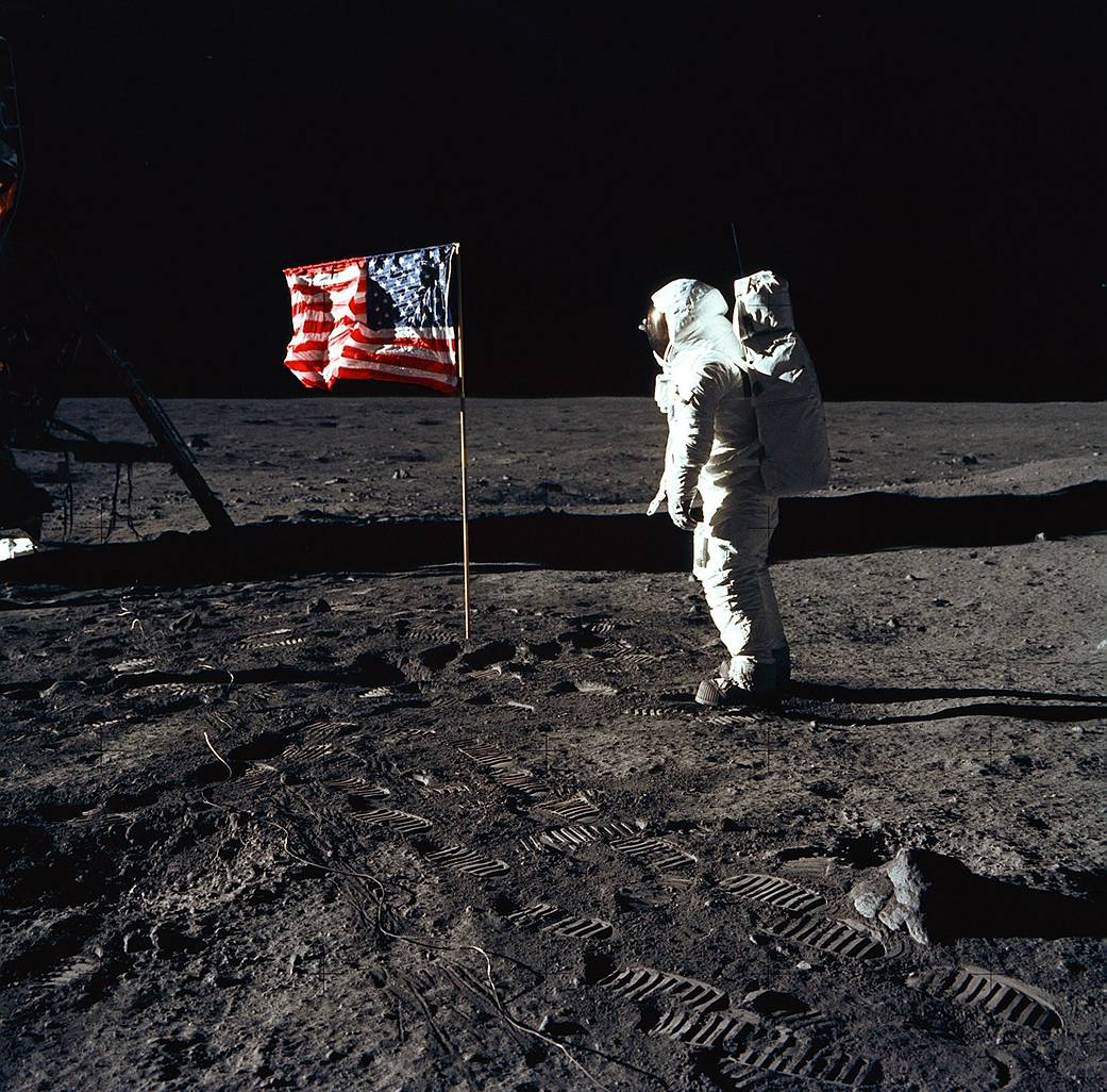 Astronaut Buzz Aldrin pozira uz zastavu Sjedinjenih Američkih Država, koju su na površinu Mjeseca astronauti postavili tijekom misije Apollo 11. Lunarni modul nalazi se na lijevoj strani fotografije, a na površini Mjeseca jasno su vidljivi otisci cipela astronauta.