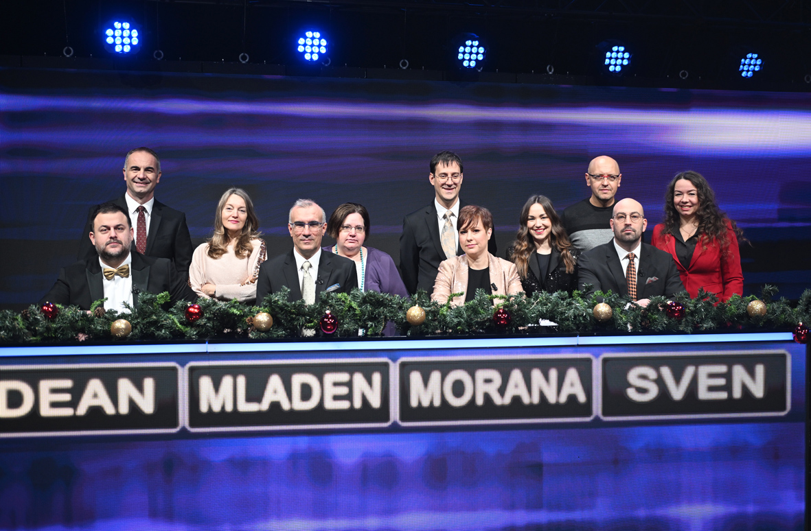 Skupina natjecatelja u svečanoj odjeći stoji iza stola ukrašenog božićnim ukrasima na televizijskom kvizu.