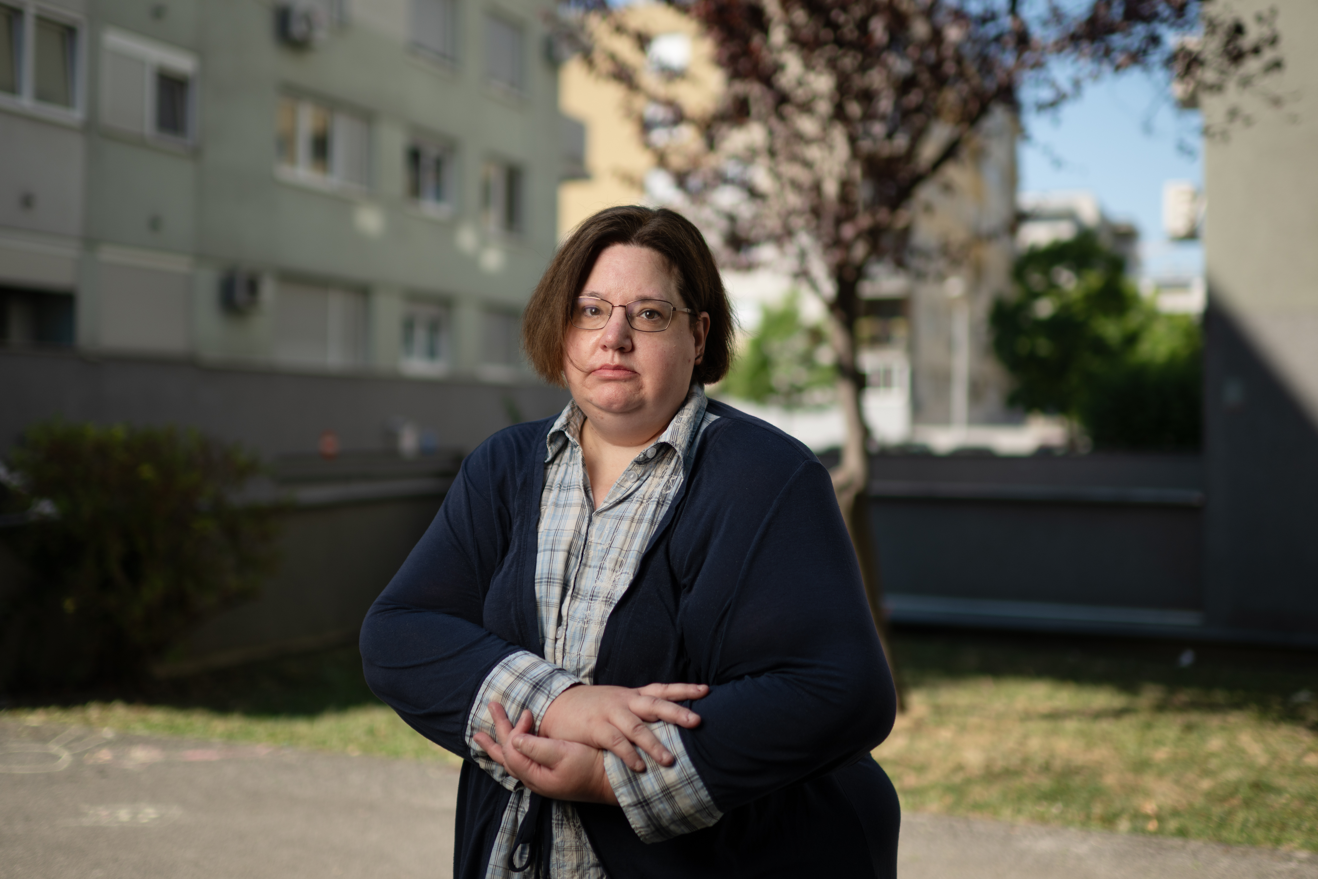 Žena s naočalama u tamnom kaputu i karirastoj košulji stoji ispred stambene zgrade s cvjetajućim drvetom.